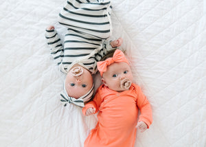 Two babies laying opposite each other on a white bed spread wearing a striped knotted gown and a coral knotted gown.