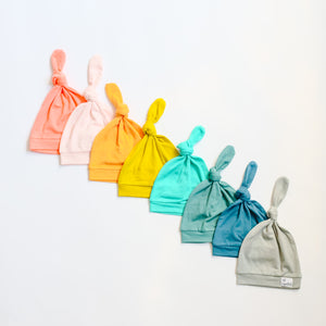 Solid color Copper Pearl top knot hats laying in rainbow order on white background